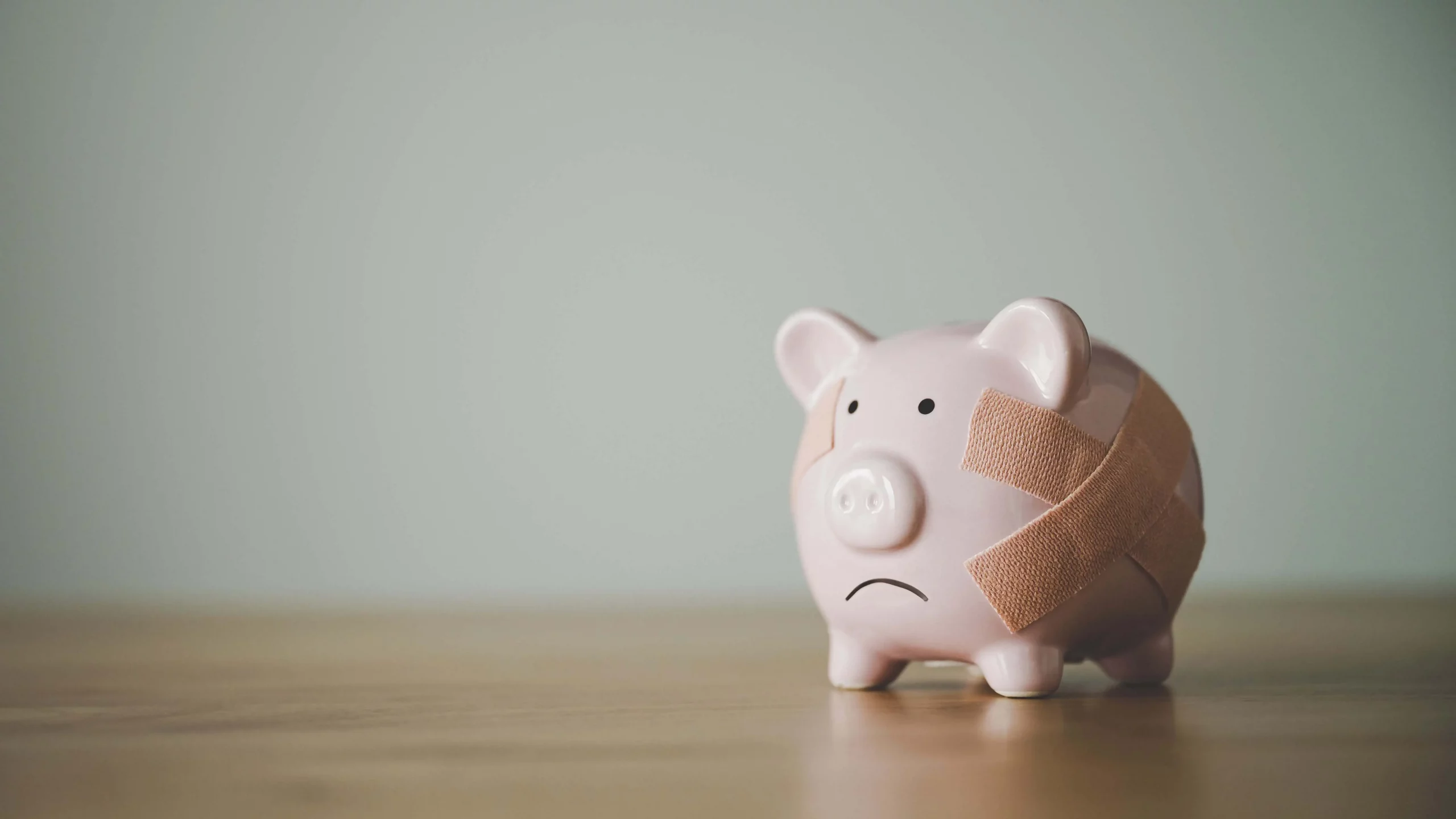 A sad piggy bank. It is rounded, and a light pink shade. It has two bandages on it's right, they are a brownish orange, and form an X. The bank is on a brown floor, and there is a grey background behind it.