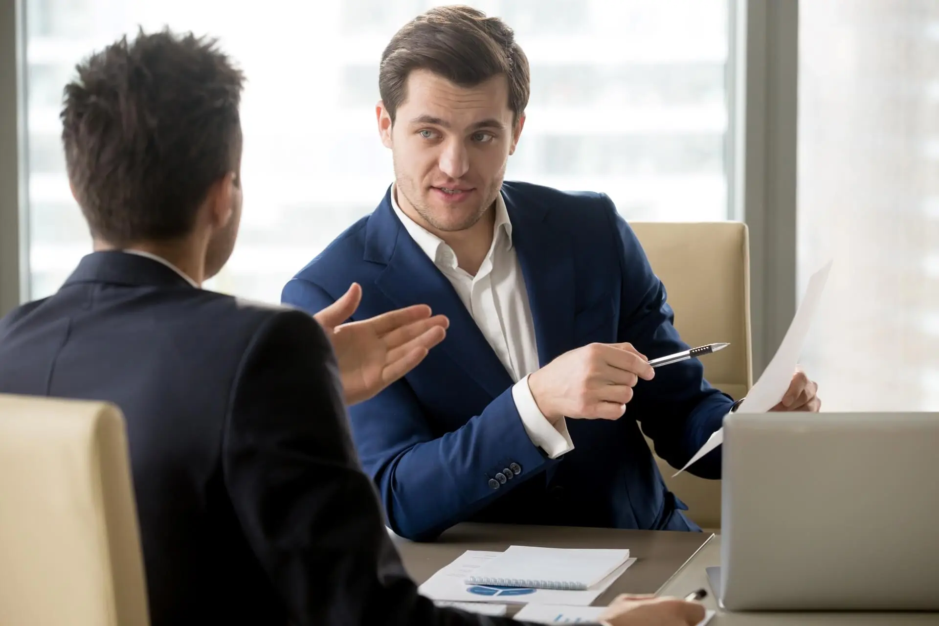 A man wearing a navy suit and a white suit sits at a desk, looking at a tax return in his hand. He's